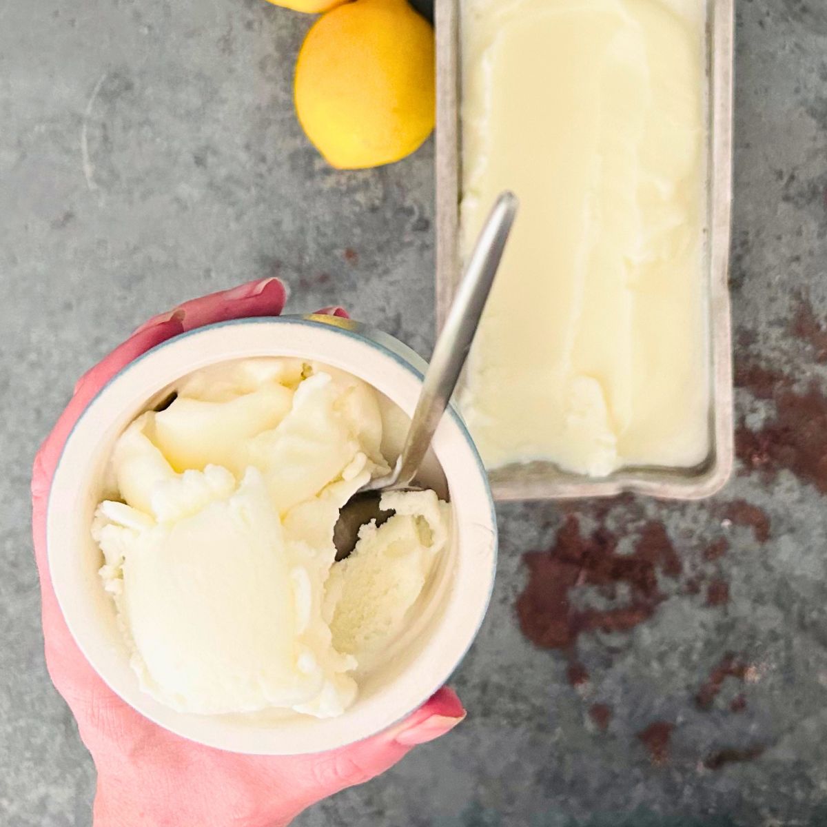 A hand is holding a small bowl of lemon ice cream with a spoon held over a a batch in loaf pan and a lemon.