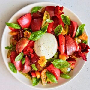Burrata and heirloom tomato salad with basil and edible flowers on a white plate.