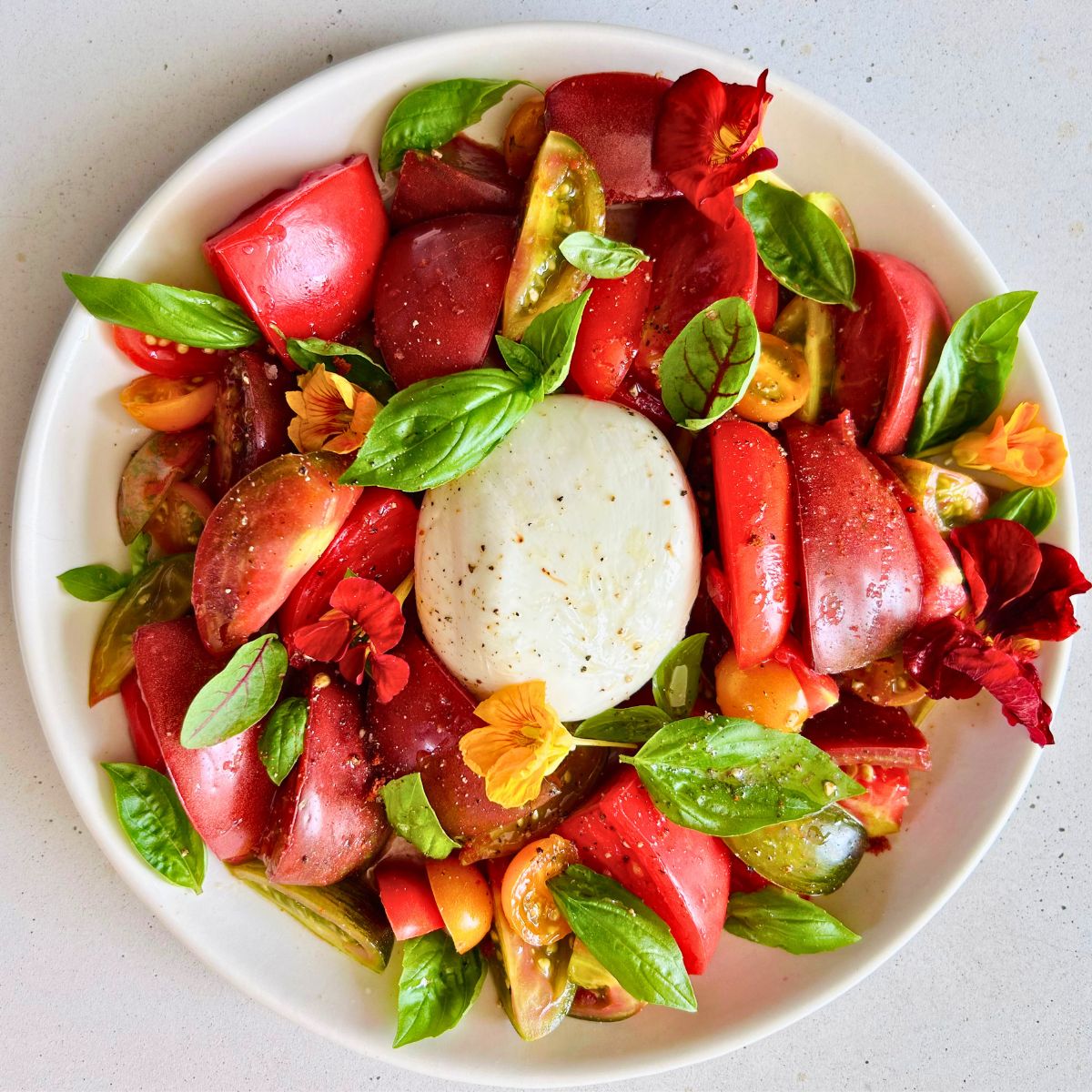 Burrata and heirloom tomato salad with basil and edible flowers on a white plate.