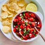 Cherry tomato pico de gallo in a white bowl with a spoon next to a plate of tortilla chips and half a lime.