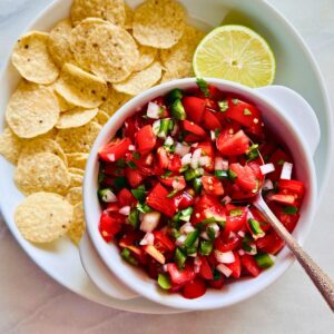 Cherry tomato pico de gallo in a white bowl with a spoon next to a plate of tortilla chips and half a lime.