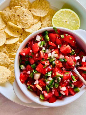 Cherry tomato pico de gallo in a white bowl with a spoon next to a plate of tortilla chips and half a lime.
