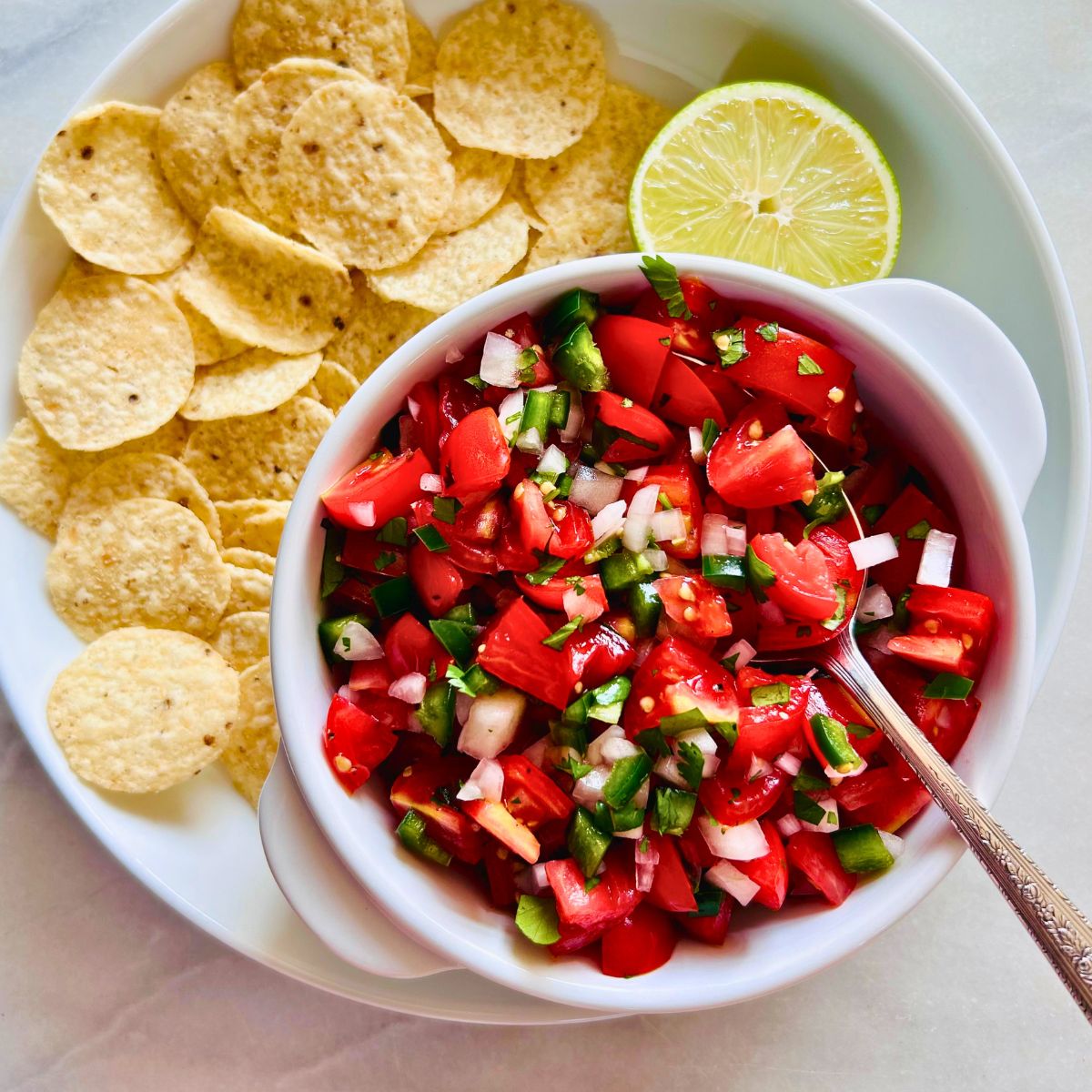 Cherry tomato pico de gallo in a white bowl with a spoon next to a plate of tortilla chips and half a lime.