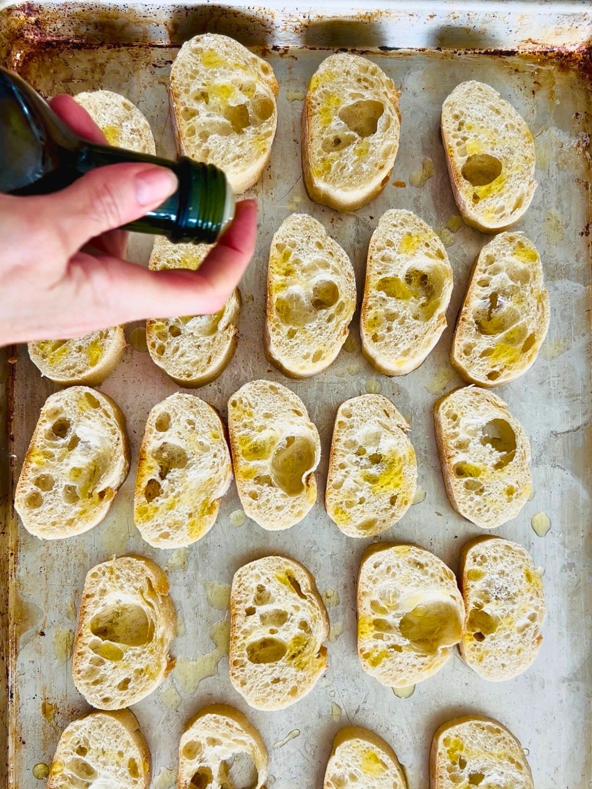 A finger is held over a bottle of olive oil while drizzling the oil over slices of bread.