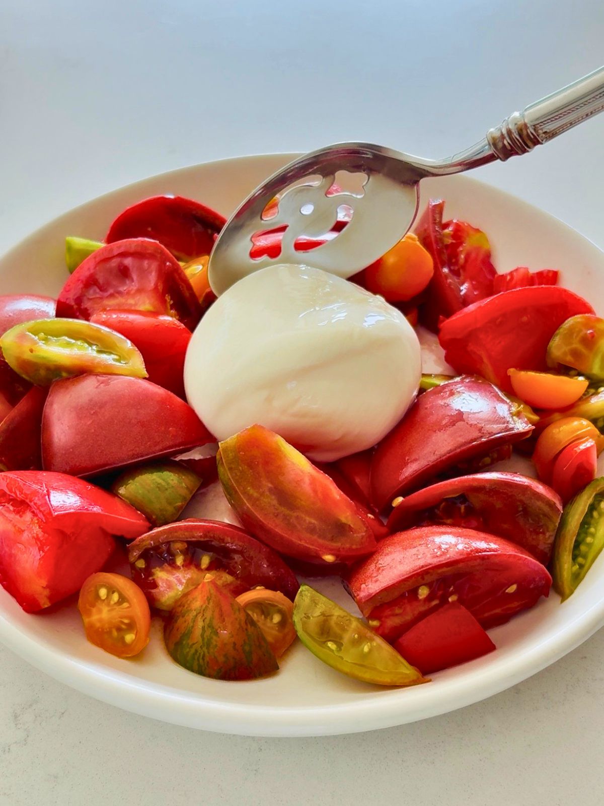 A slotted spoon is placing burrata in the center of a plate of salted heirloom tomatoes from the garden.