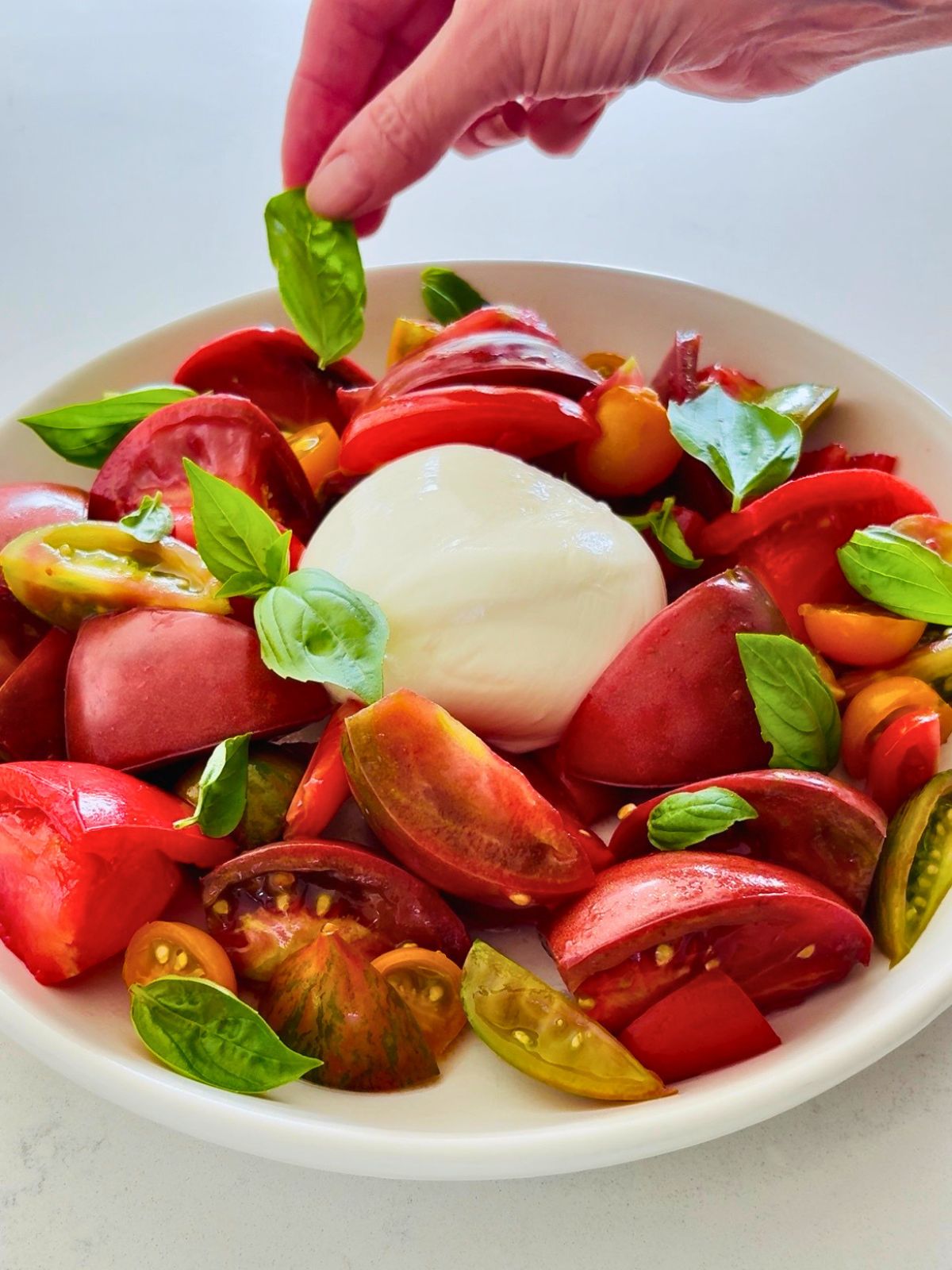 A hand is placing a basil leaf onto a plate of burrata and heirloom tomato salad.