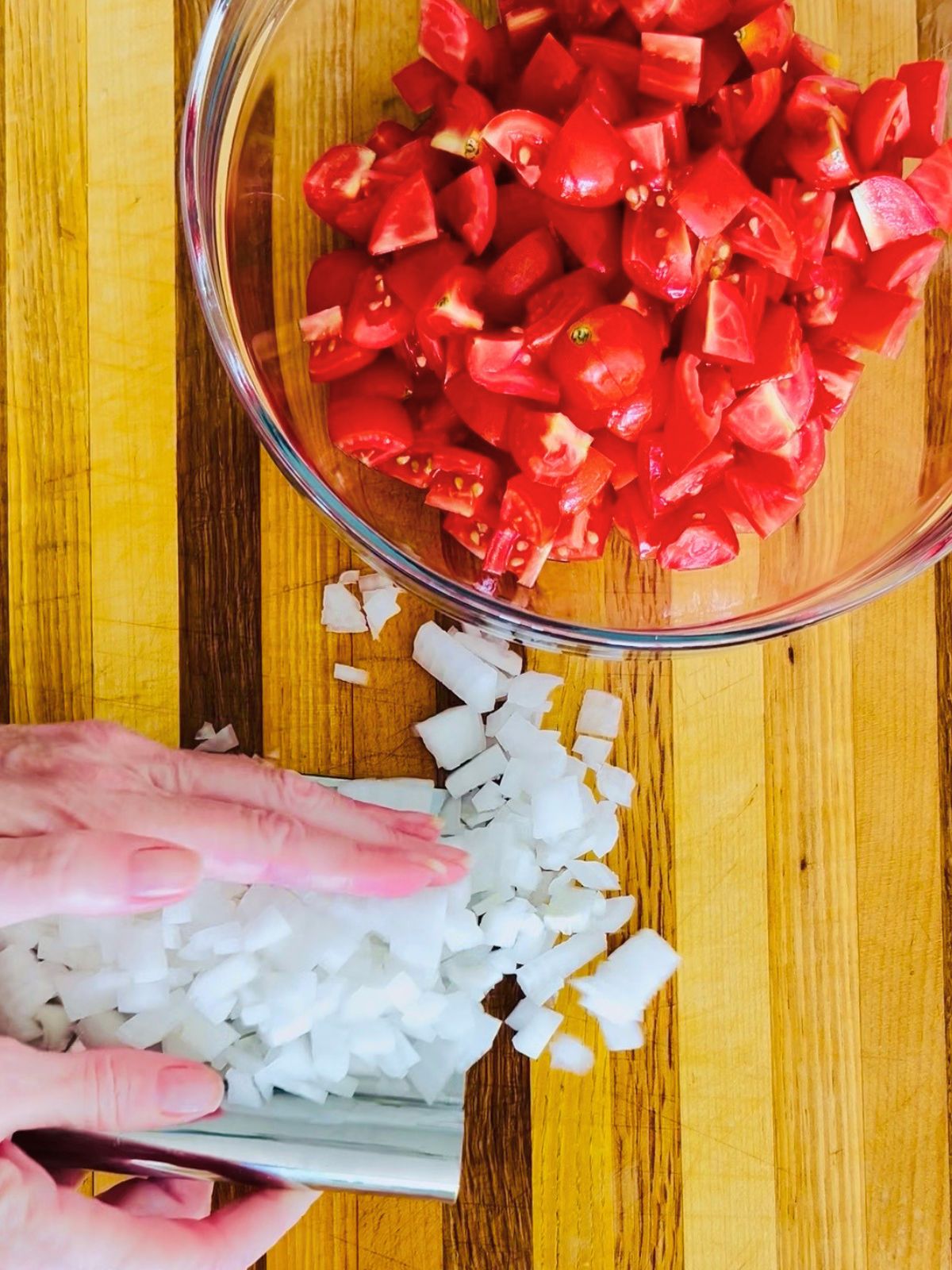 A bench scraper is scooping up diced white onions to a bowl of chopped cherry tomatoes.
