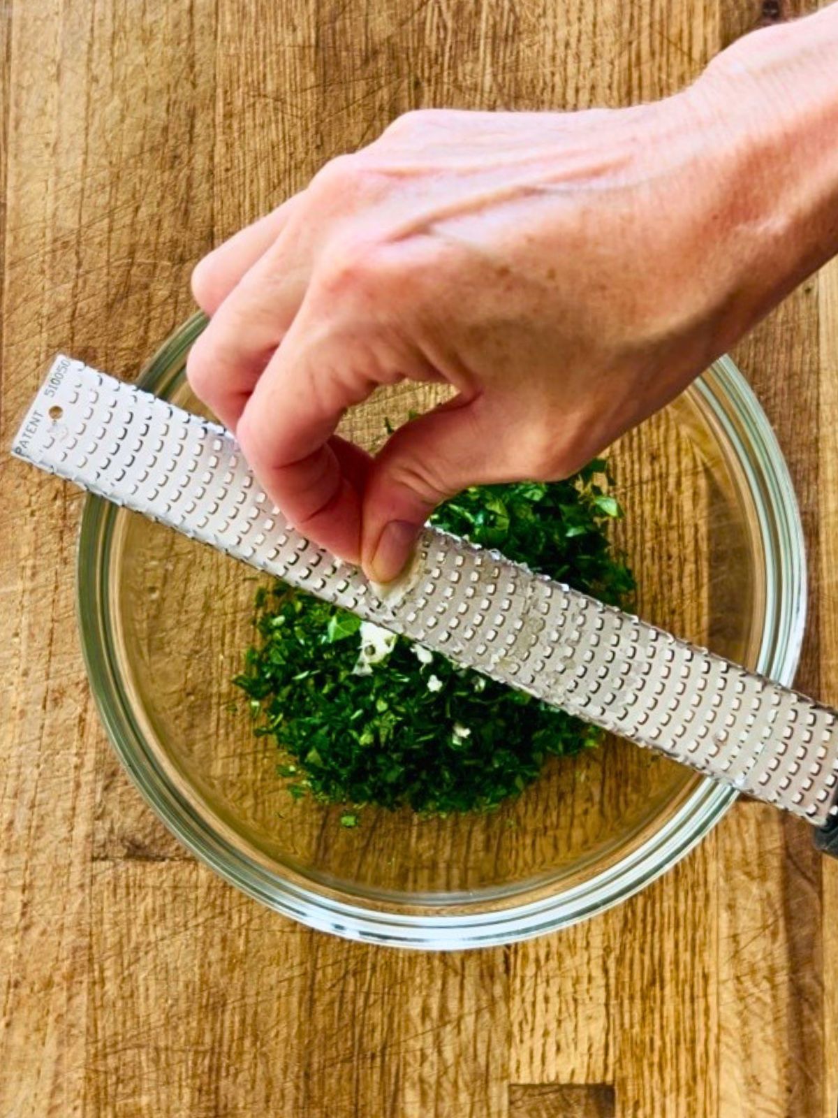 A garlic clove is grated with a Microplane rasp set over a bowl of chopped herbs.