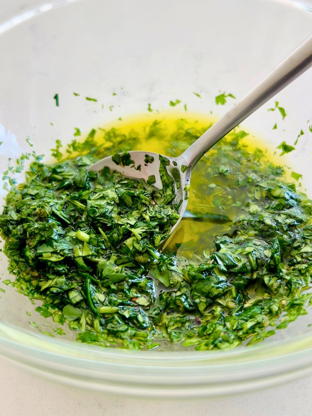 A spoon is stirring olive oil into a glass bowl of chimichurri sauce.