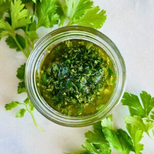 Chimichurri made with cilantro and parsley in a small mason jar next to sprigs of herbs.