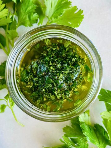 Chimichurri made with cilantro and parsley in a small mason jar next to sprigs of herbs.