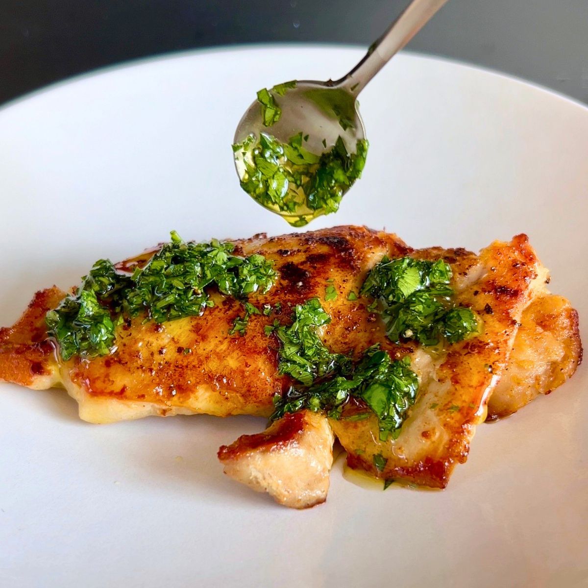 Chimichurri sauce with cilantro and parsley is spooned over a seared skinless chicken thigh on a white plate.