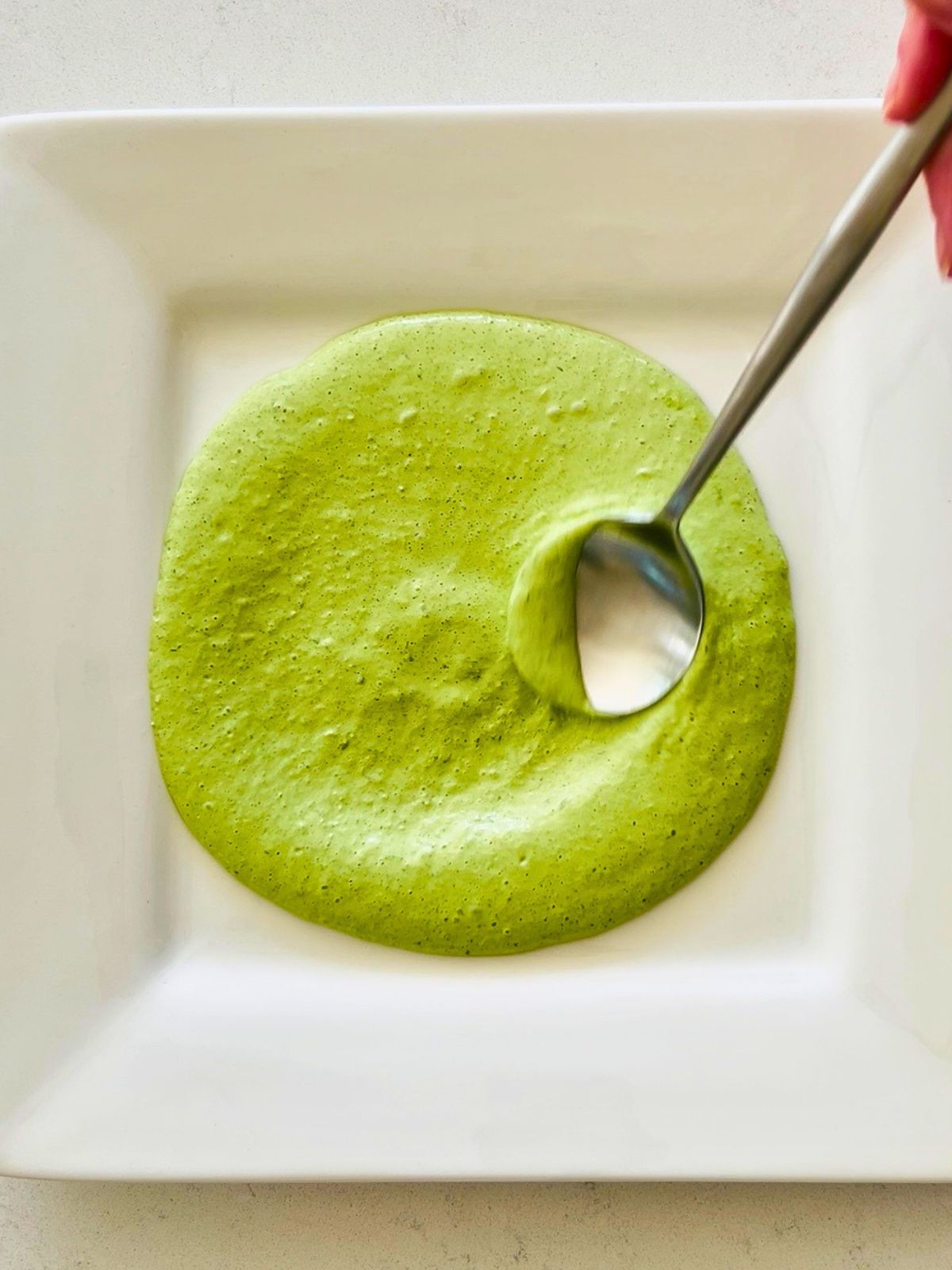 A spoon spreading green goddess around a square white plate.