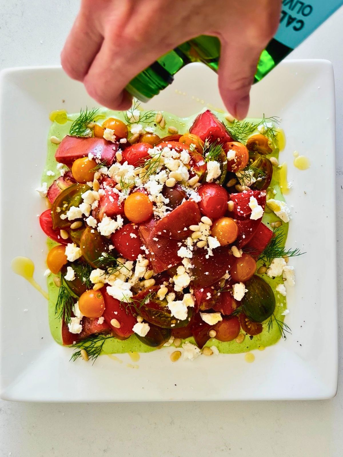 Olive oil is drizzling over a white plate of tomatoes, feta, pine nuts, dill, and green goddess dressing.