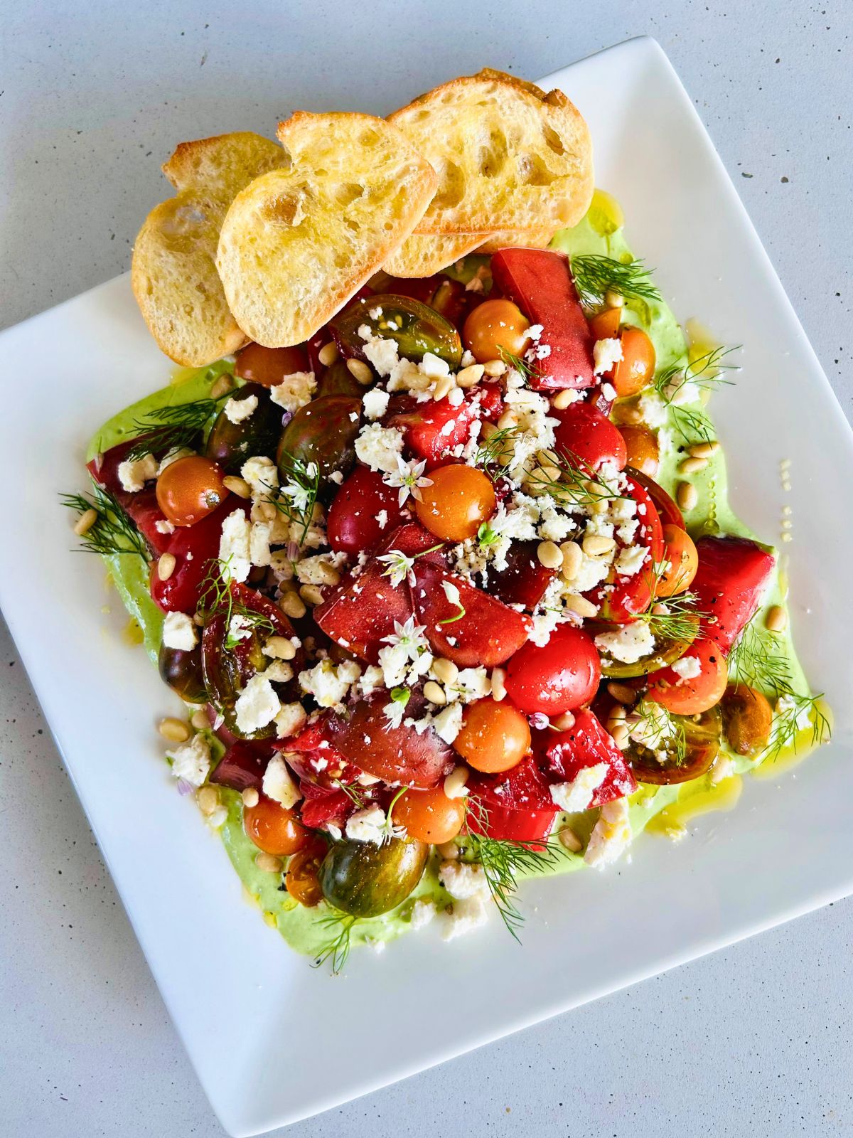 A white plate of fresh tomato salad over green goddess dressing with toasted sliced baguette.