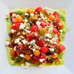 A white plate with fresh tomato salad with feta, pine nuts, dill, and green goddess dressing.