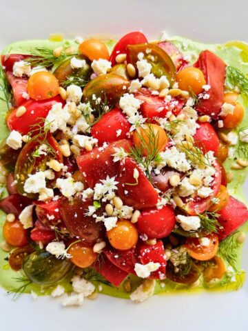 A white plate with fresh tomato salad with feta, pine nuts, dill, and green goddess dressing.