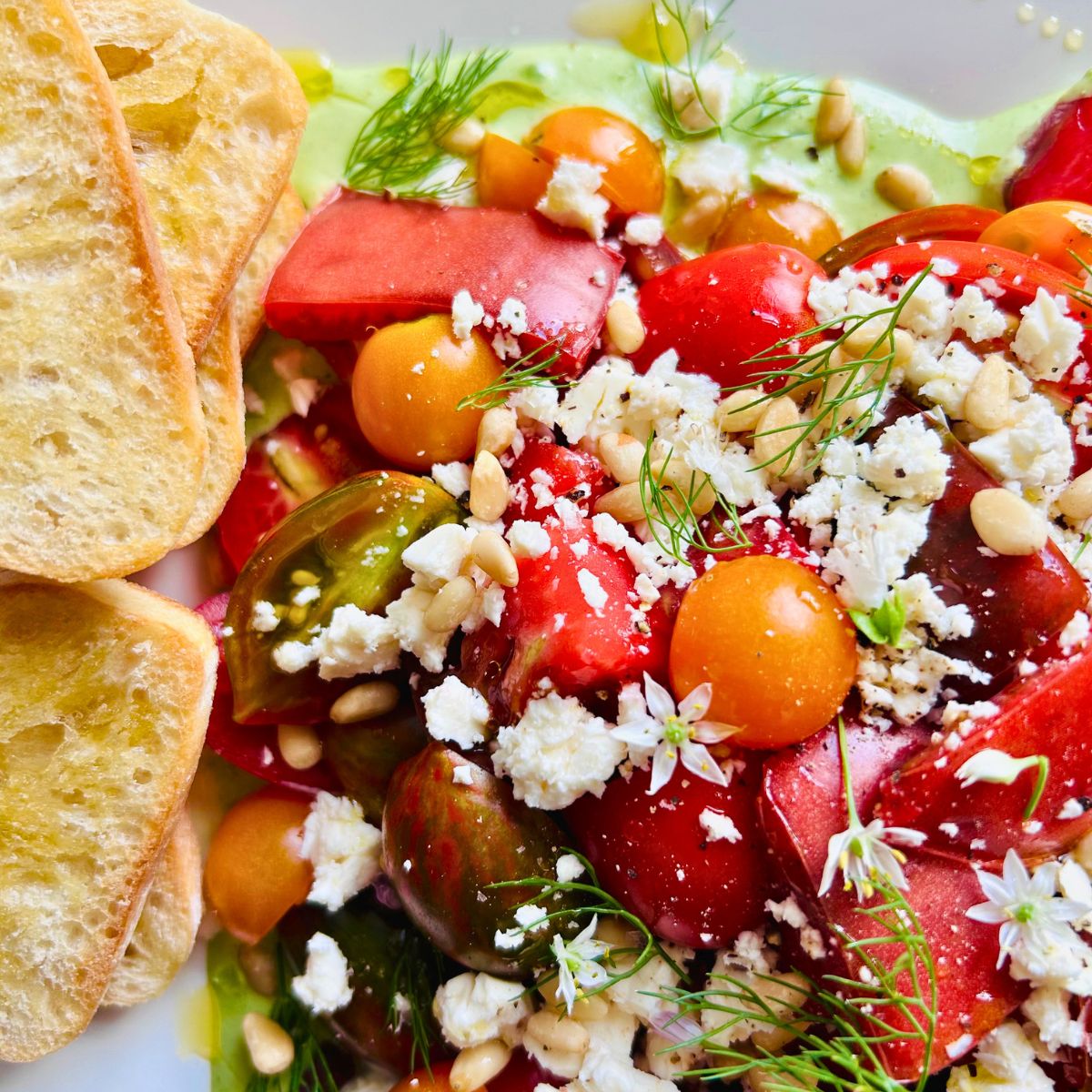 Toasted sliced bread next to a plate of fresh tomato salad with feta, toasted pine nuts, and green goddess dressing.