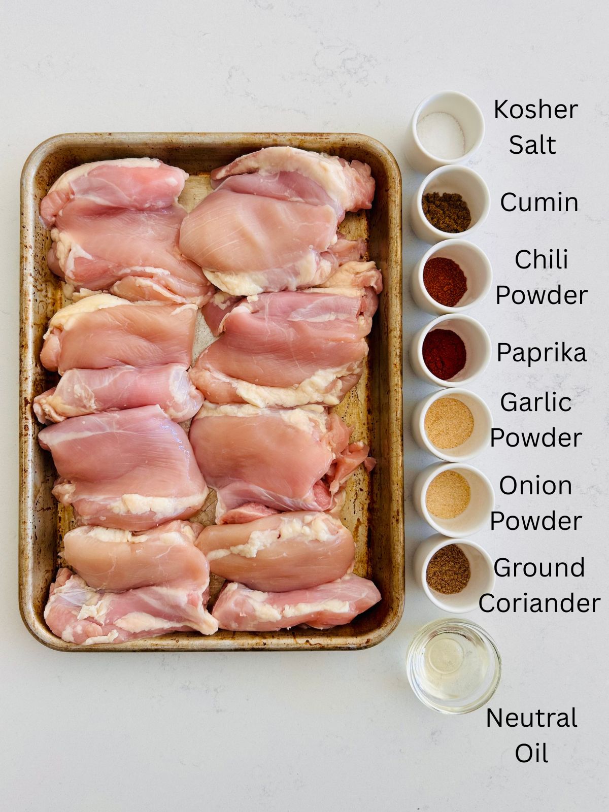 Boneless and skinless chicken thighs on a rimmed baking sheet next to small bowls of pantry spices needed to make taco seasoning.