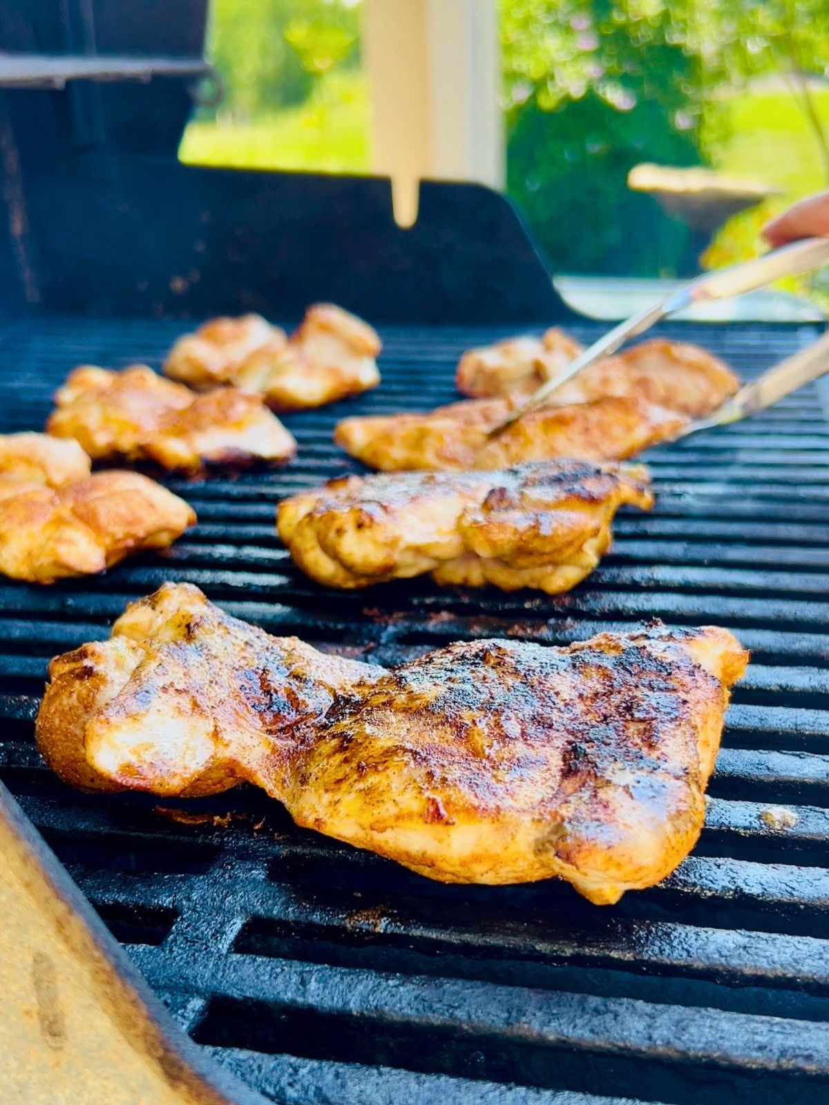 Boneless, skinless chicken thighs are getting flipped with tongs on gas grill grates.