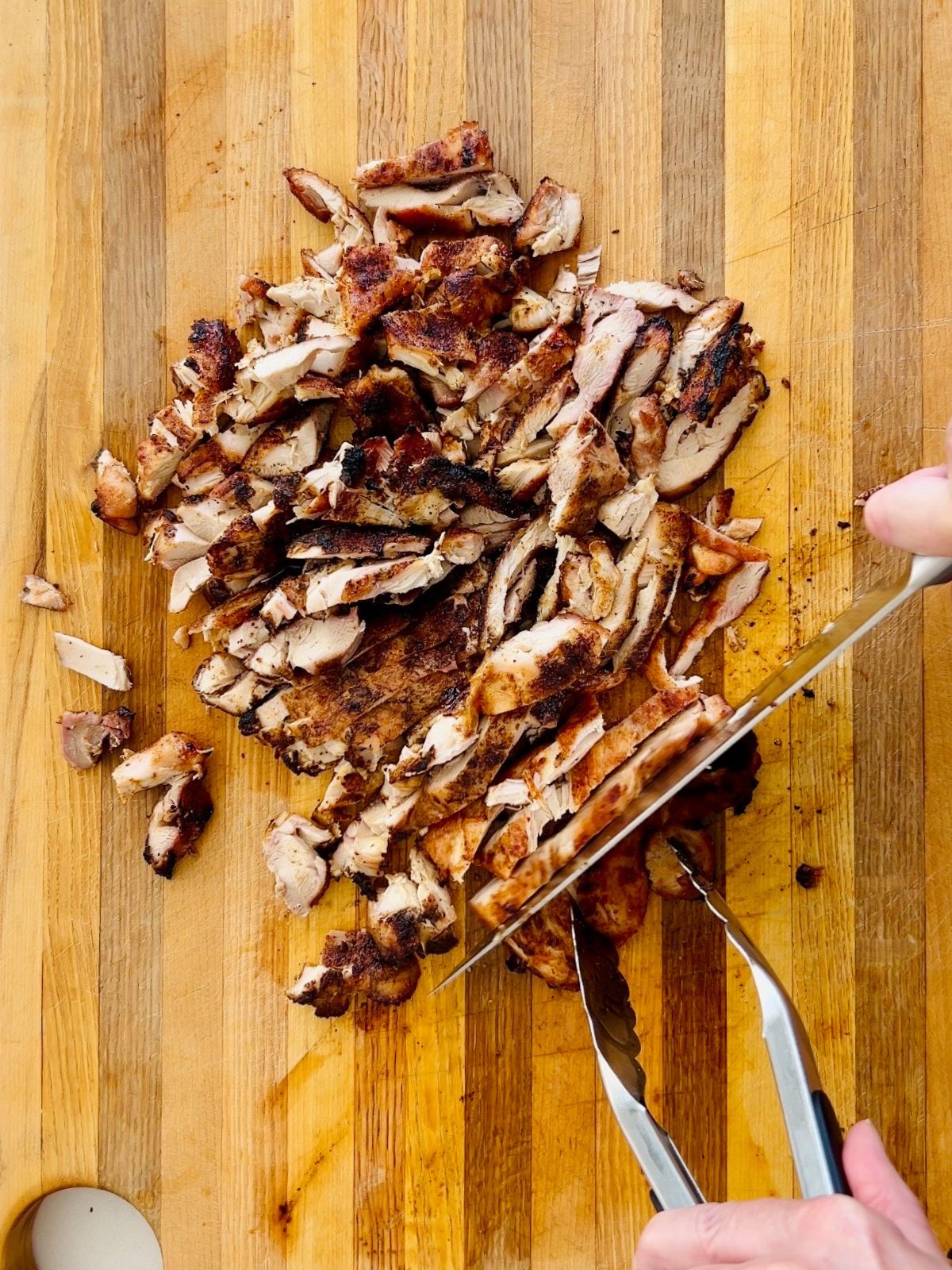 Grilled boneless, skinless chicken thighs are chopped by a knife into bite-sized pieces over a wooden cutting board.