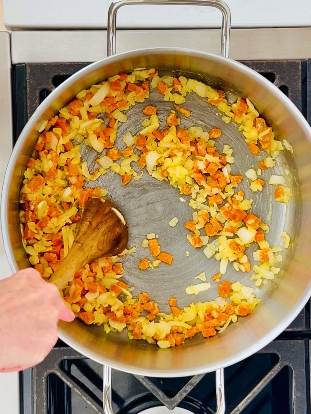 A wooden spoon is used to stir flour into sautéed carrots, onions, and garlic in a large pot.