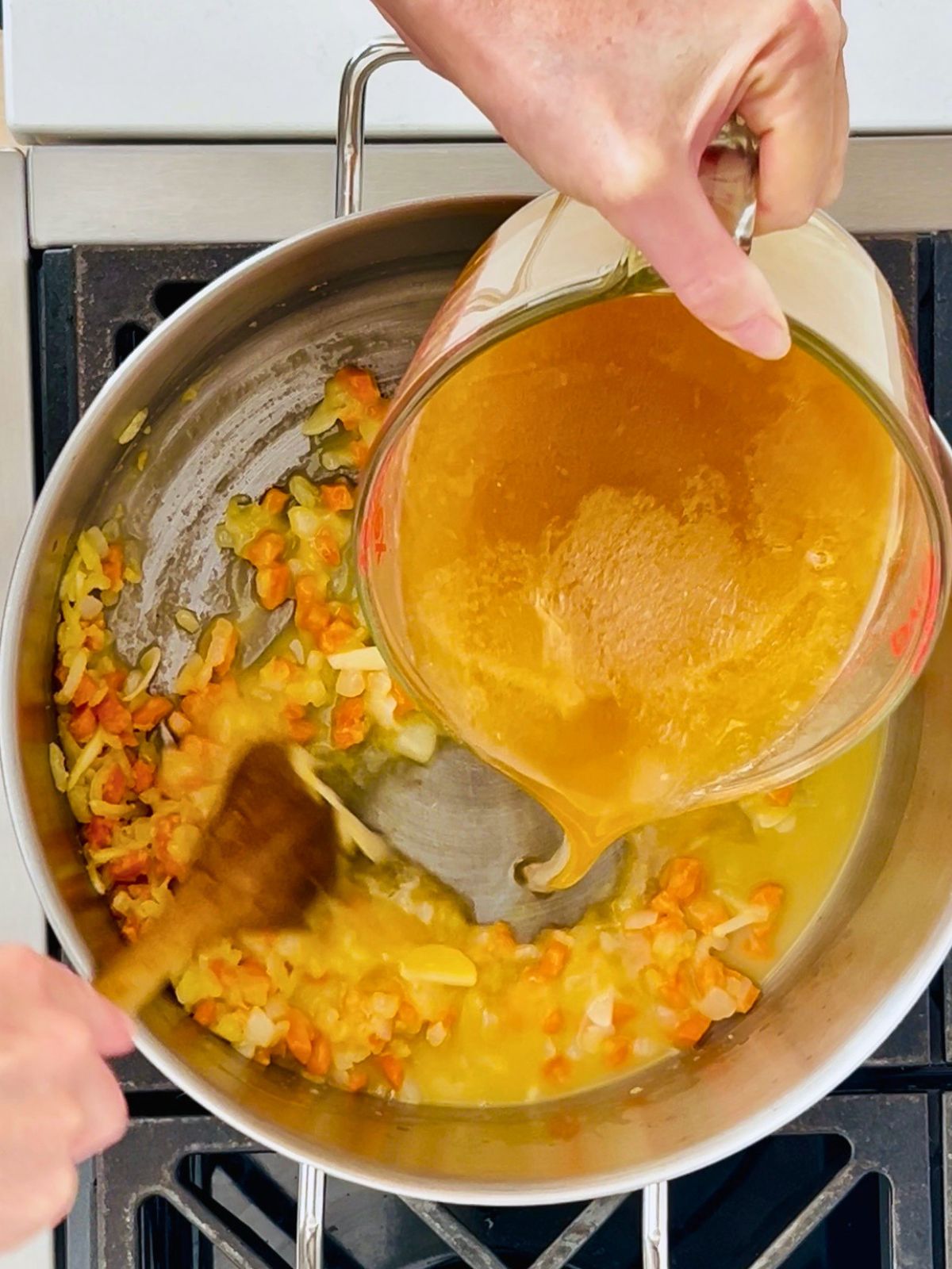 Brown chicken stock is slowly added while stirring into the carrot, onion, and roux in a stock pot.