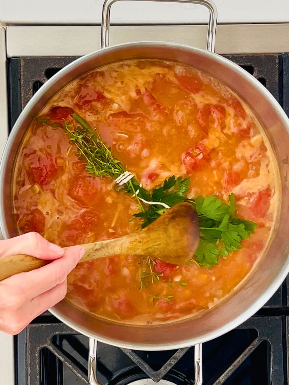A bundle of fresh herbs is added to a pot of whole tomatoes and chicken broth for tomato soup.