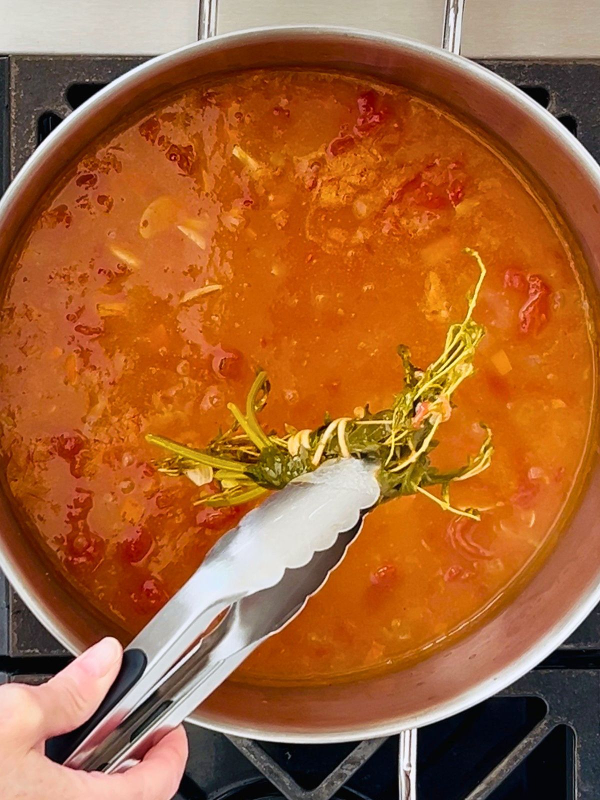 Tongs are removing a bundle of simmered herbs from a pot of tomato soup before blending.