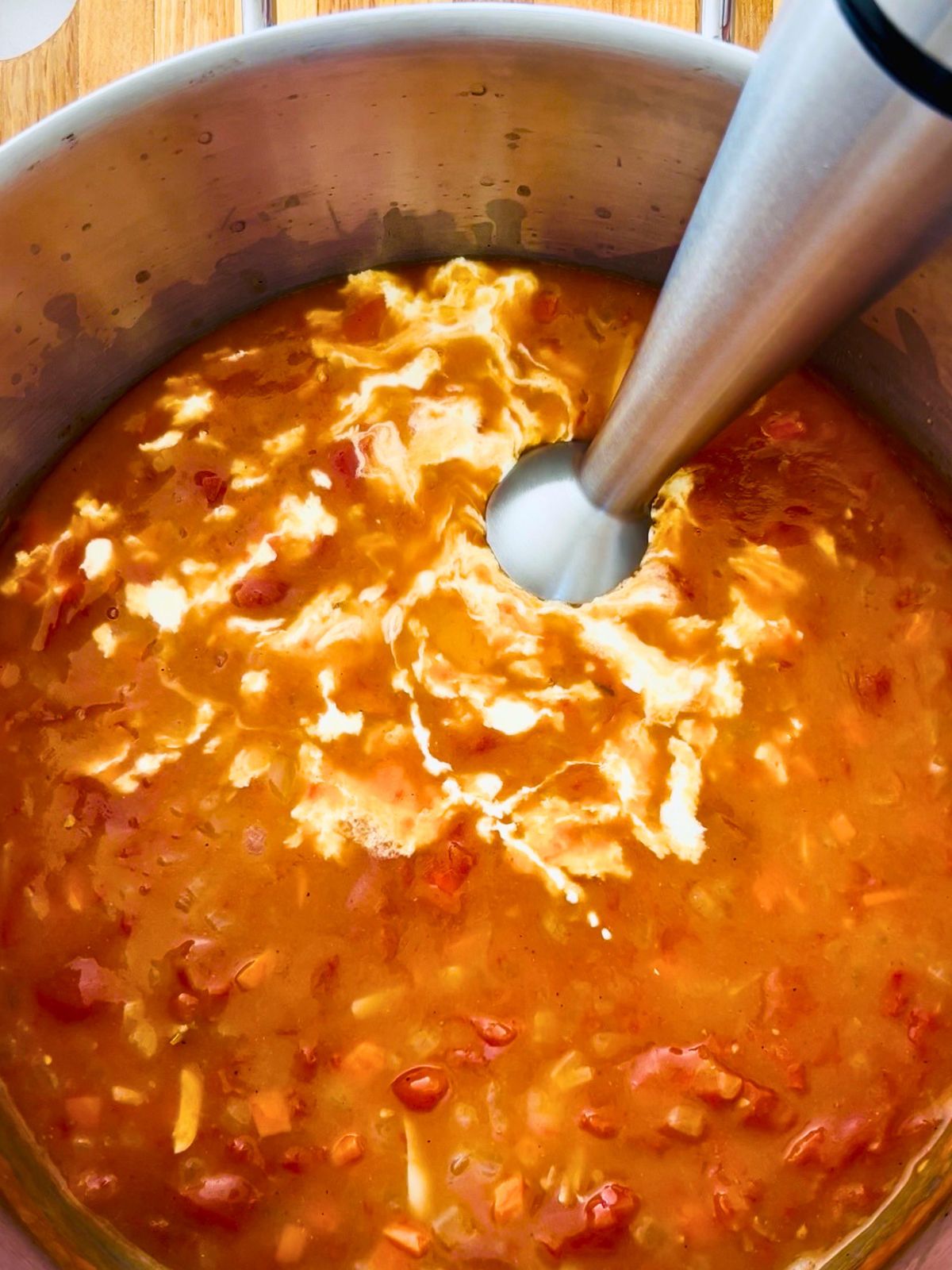 A pot of cooked canned tomatoes, chicken stock, vegetables, and cream are blended with an immersion blender to make creamy tomato soup.