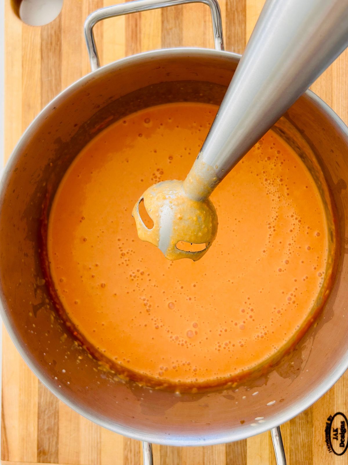 The stick of an immersion blender held over a pot of blended creamy tomato soup.