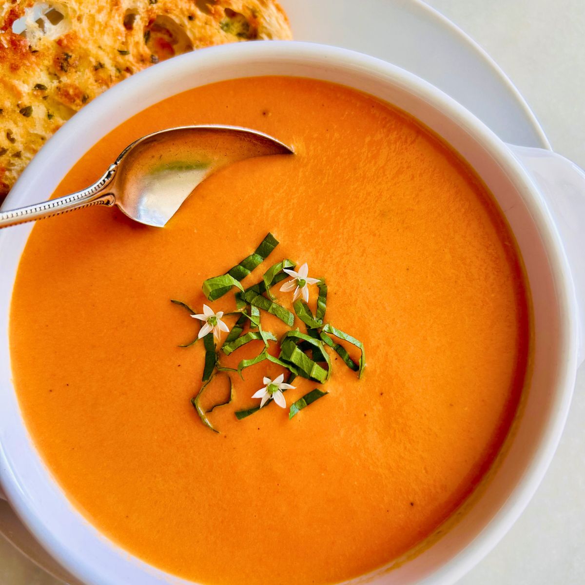 A silver spoon is going into a white bowl of the best creamy tomato soup garnished with basil chiffonade and chive flowers.