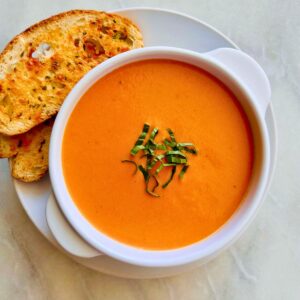 Creamy tomato soup topped with sliced basil leaves in a white bowl resting on a white plate with cheesy sliced bread to the side.