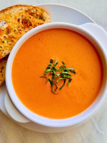 Creamy tomato soup topped with sliced basil leaves in a white bowl resting on a white plate with cheesy sliced bread to the side.