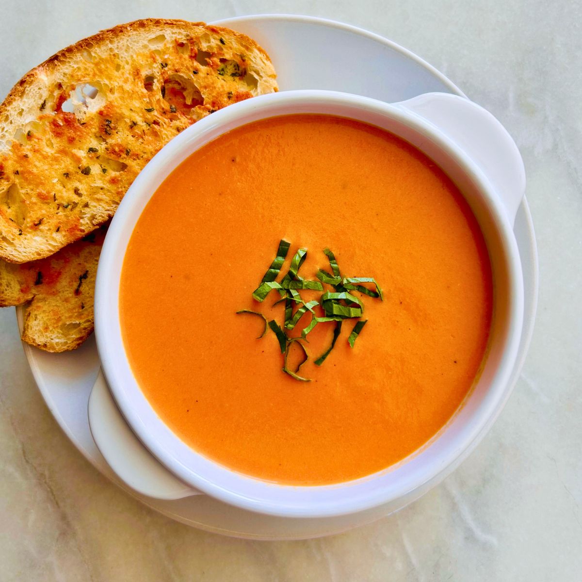 Creamy tomato soup topped with sliced basil leaves in a white bowl resting on a white plate with cheesy sliced bread to the side.