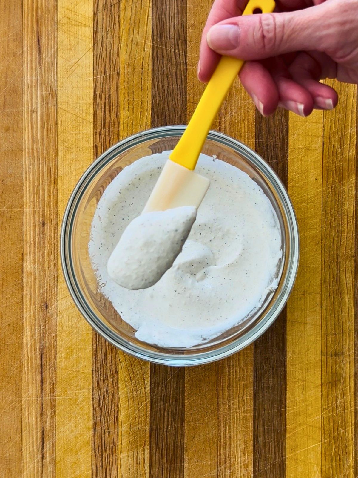 A small rubber spatula is holding creamy horseradish sauce over a glass bowl set on a cutting board.