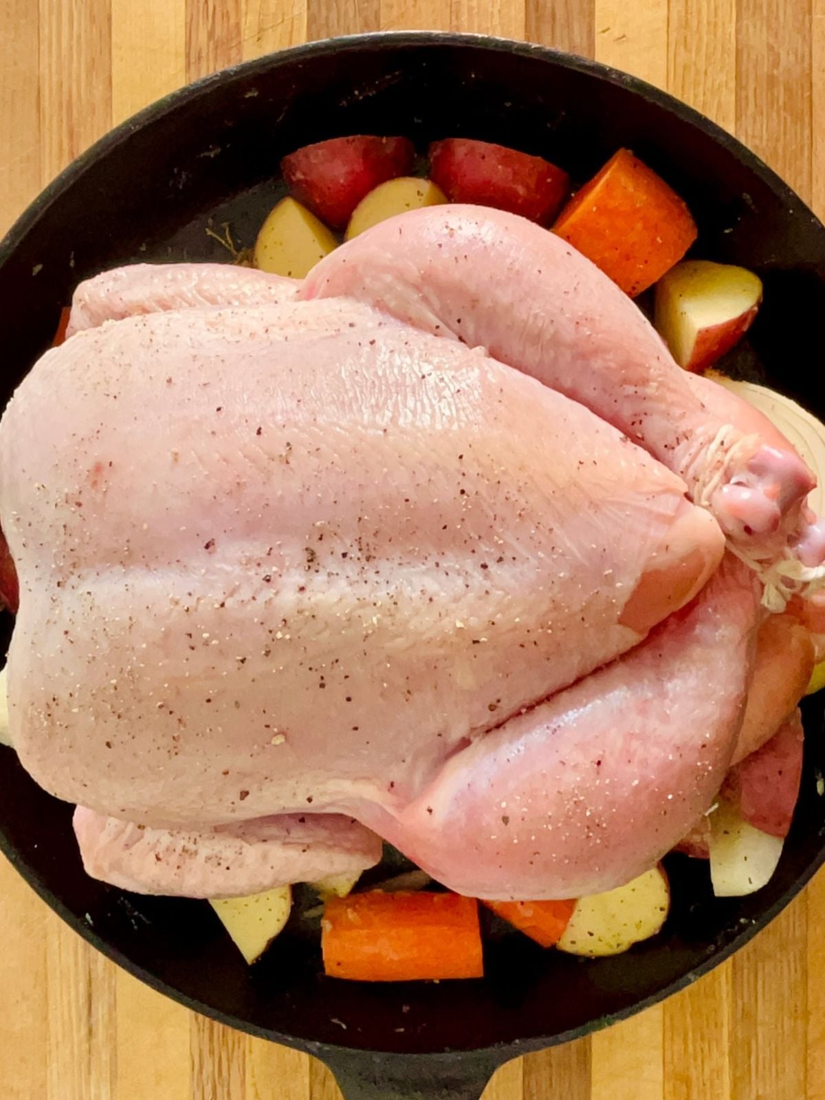 A dry-brined uncooked whole chicken is resting on top of root vegetables in a cast iron skillet.