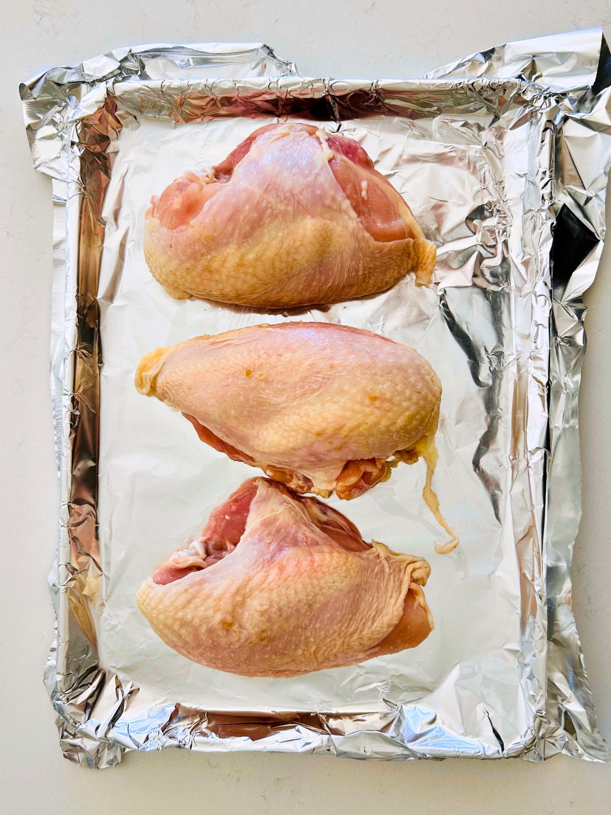 Three bone-in, skin-on chicken breasts on a foil lined rimmed baking sheet.