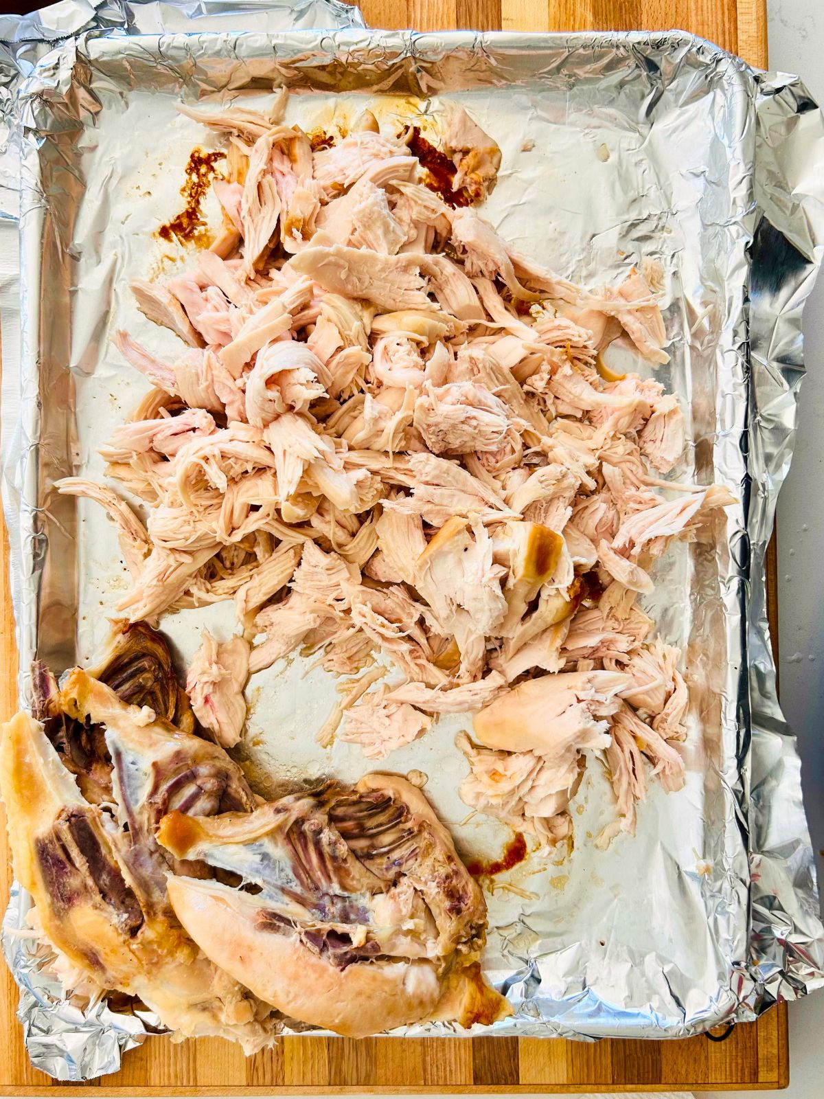 Shredded pieces of chicken breast with their bones in the corner of a foil-lined rimmed baking sheet.