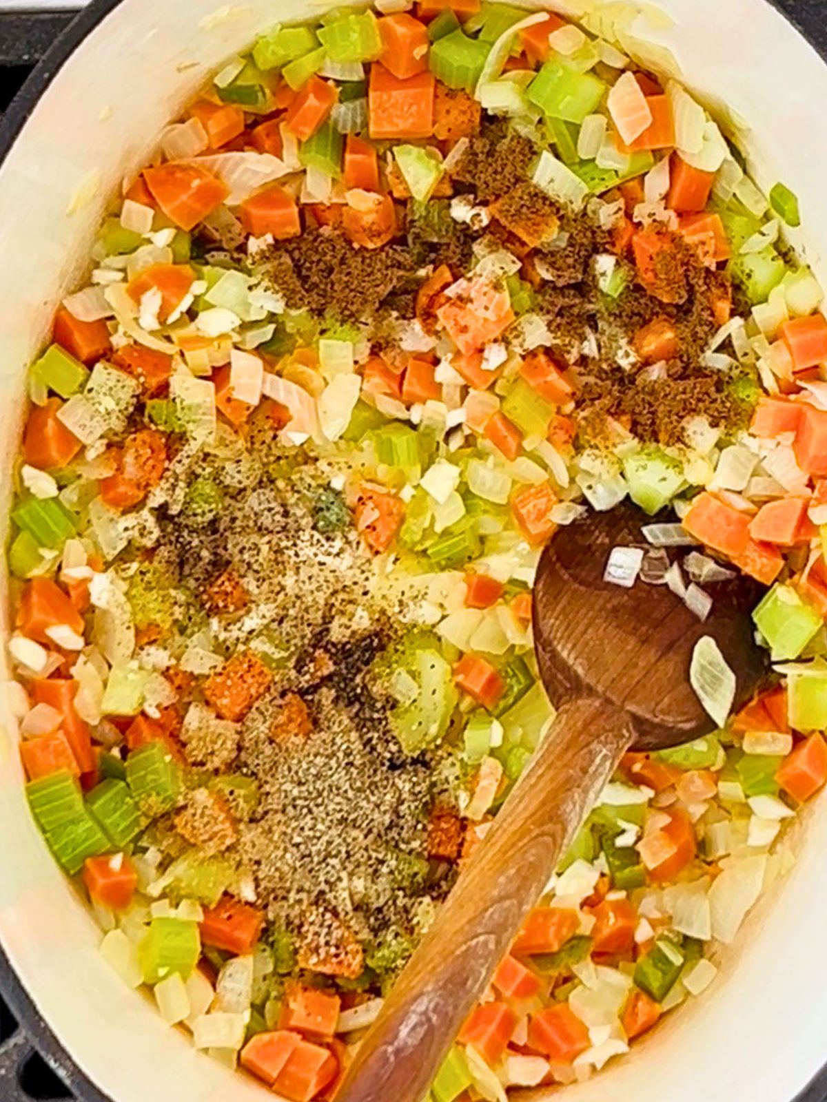 A wooden spoon is stirring cumin, coriander, and black pepper and cooked mirepoix in a pot.