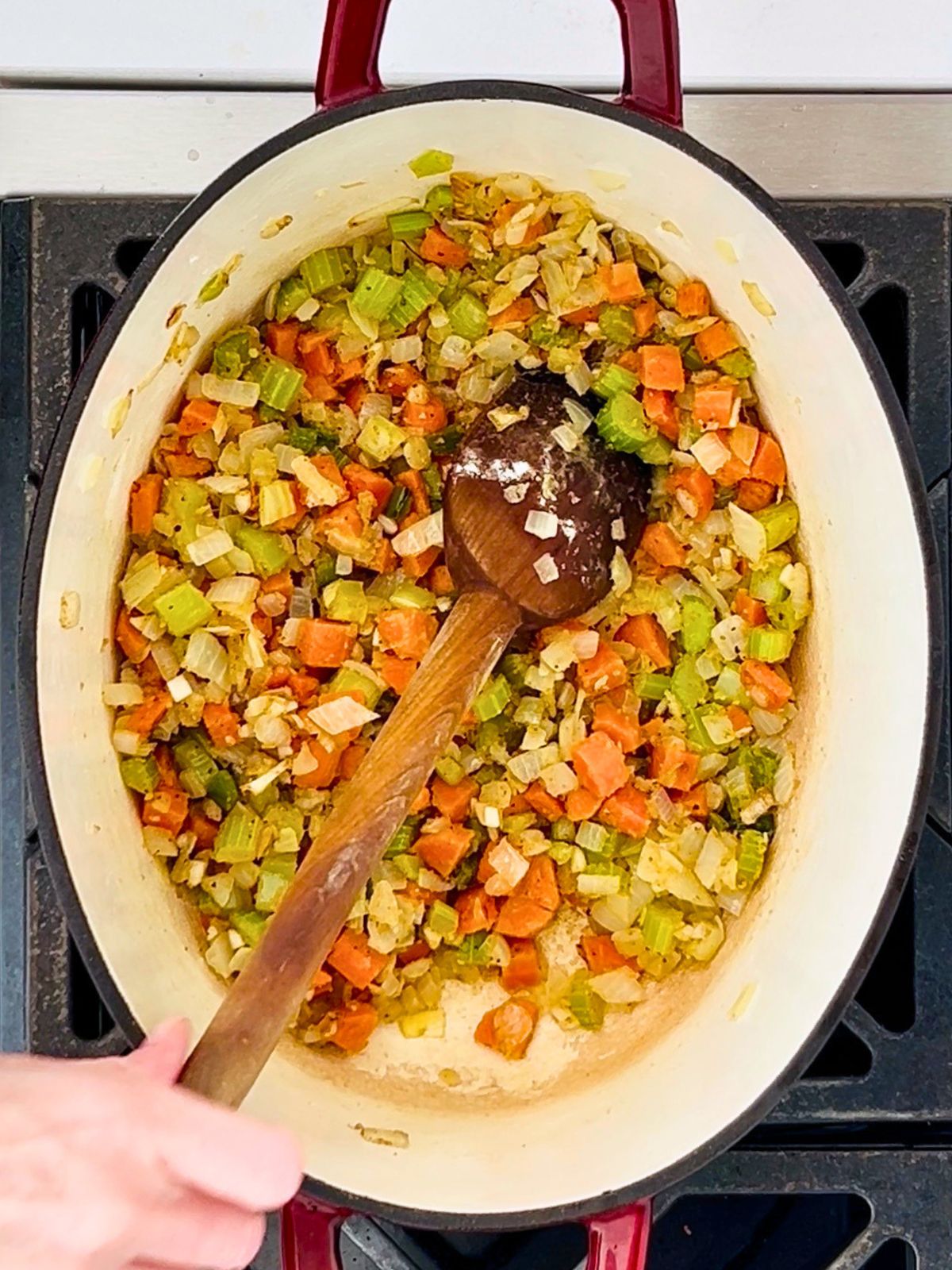A wooden spoon is stirring flour into a pot of softened onions, celery, carrots, and jalapenos over a burner.