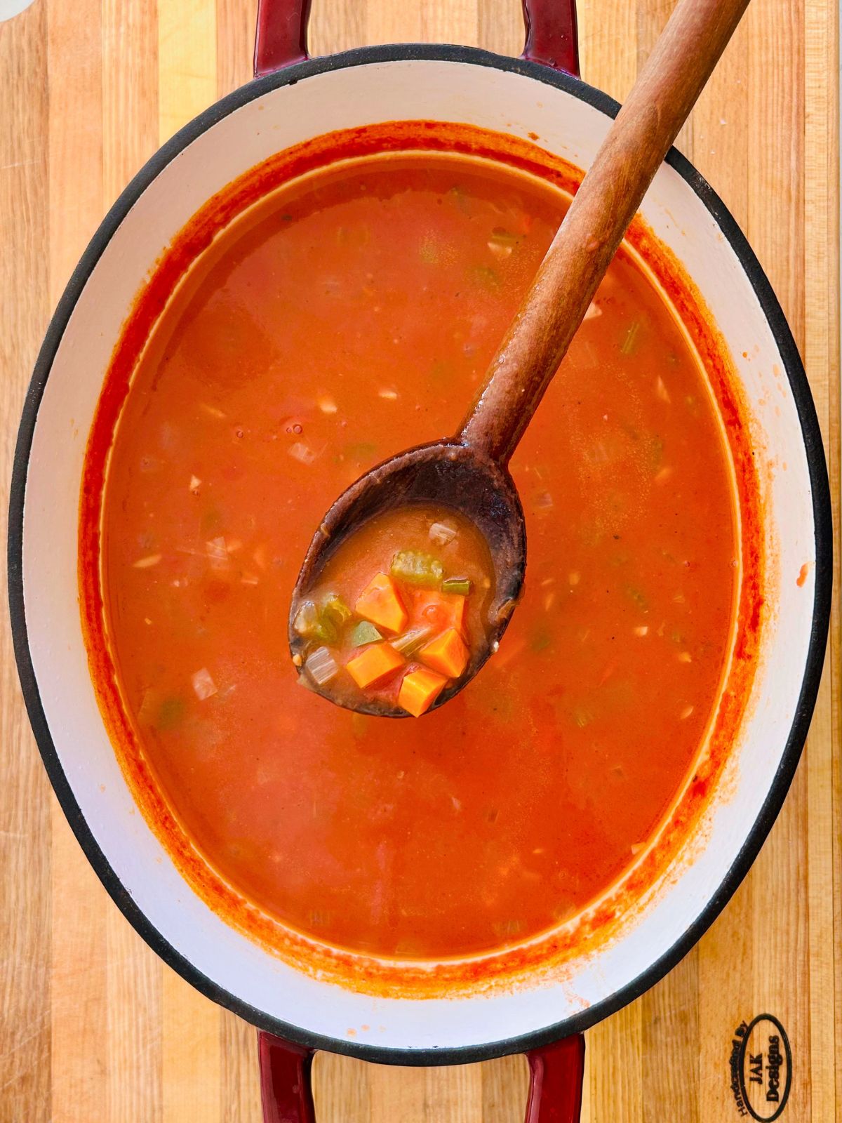 A wooden spoon holding cooked vegetables and tomato broth over a pot of Mexican soup resting on top of a cutting board.