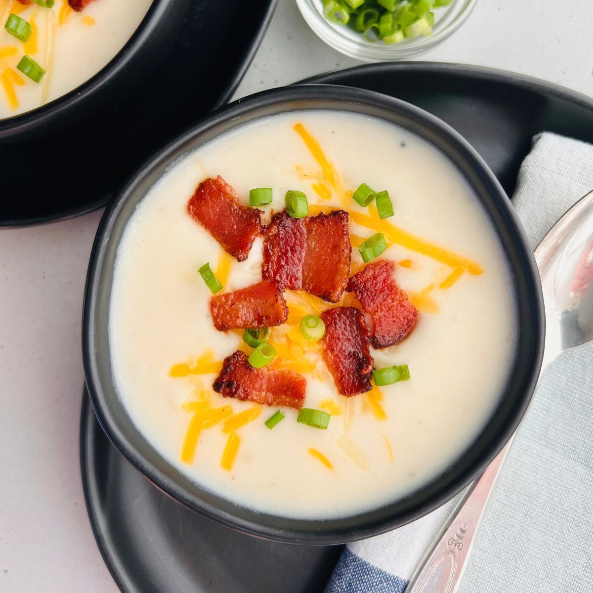 Copycat Panera baked potato soup in a black bowl on top of a black plate next to a silver spoon and napkin.