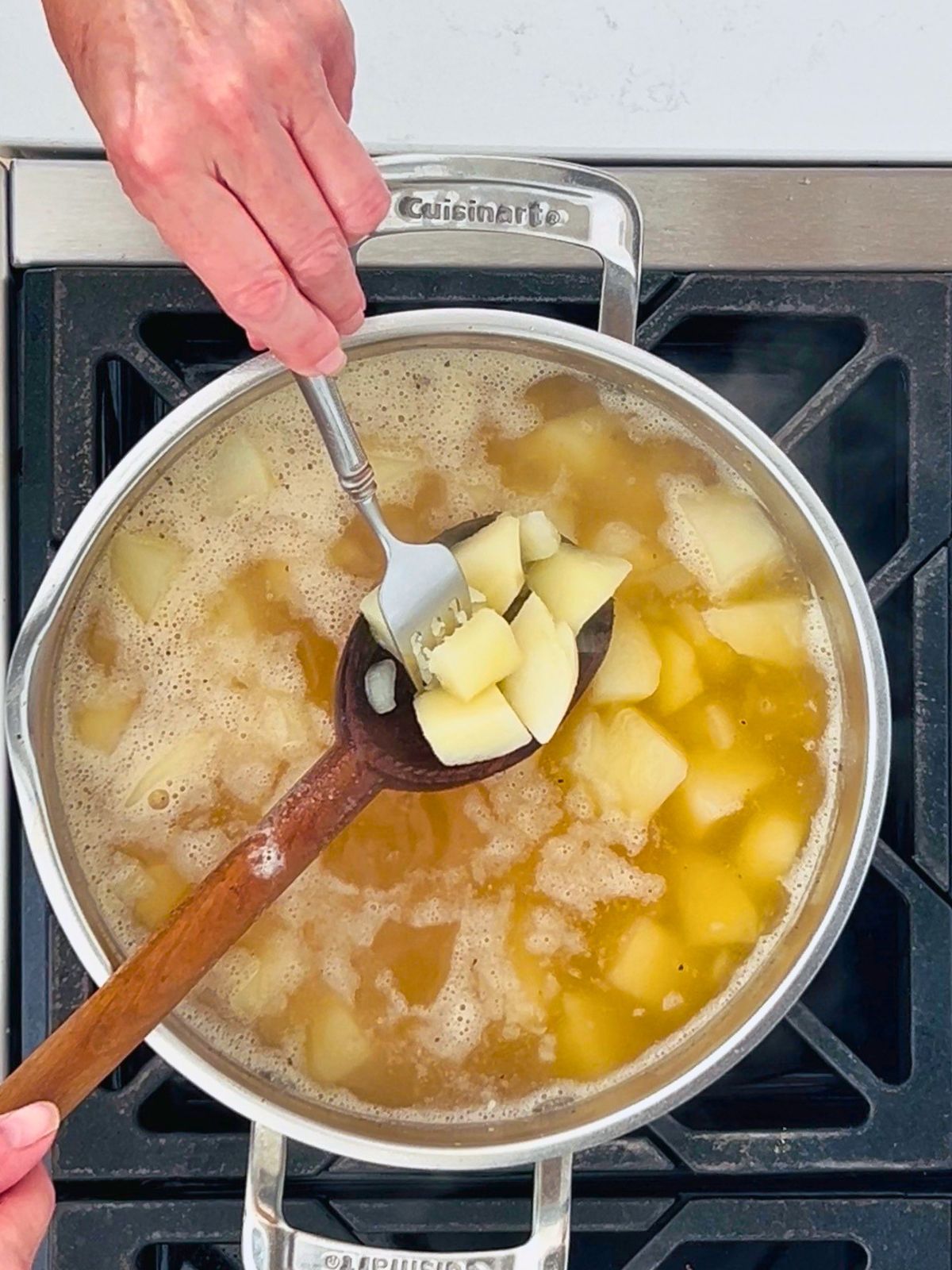 A fork is piercing a cooked piece of potato held in a wooden spoon over a simmering pot of chicken broth and potatoes.