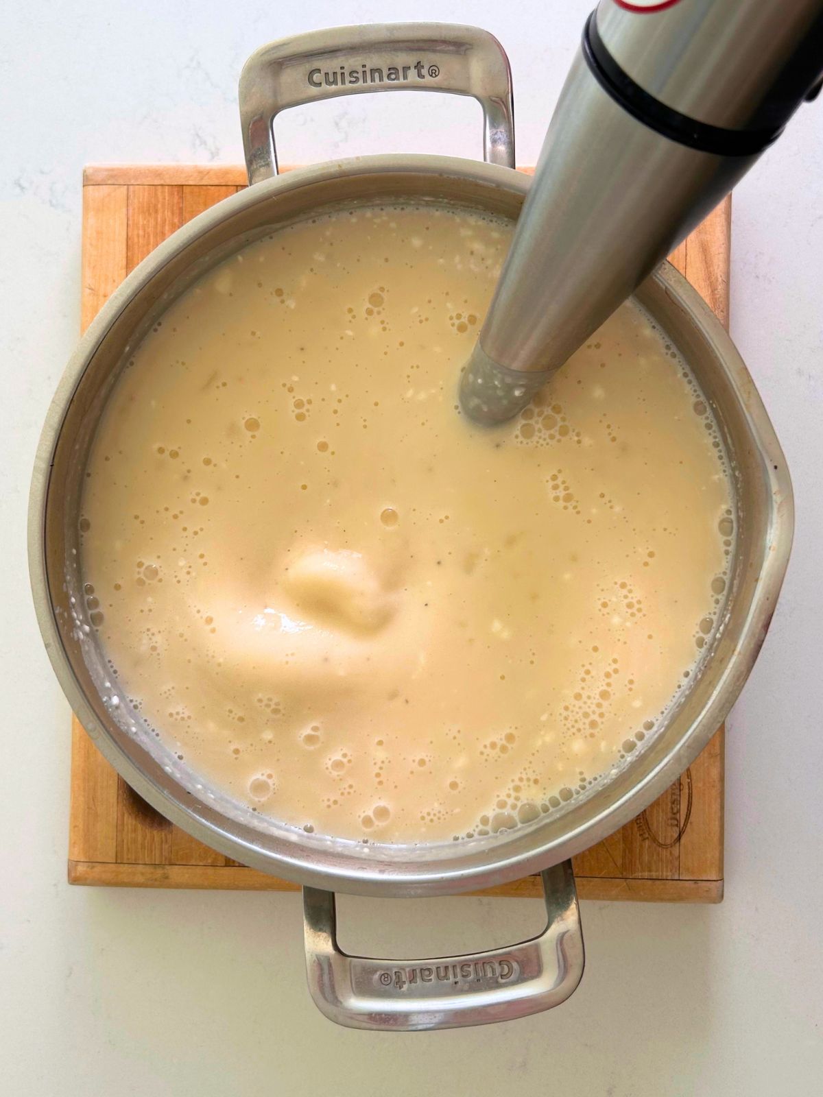 An immersion blender is blending cream cheese into a pot of potato soup.
