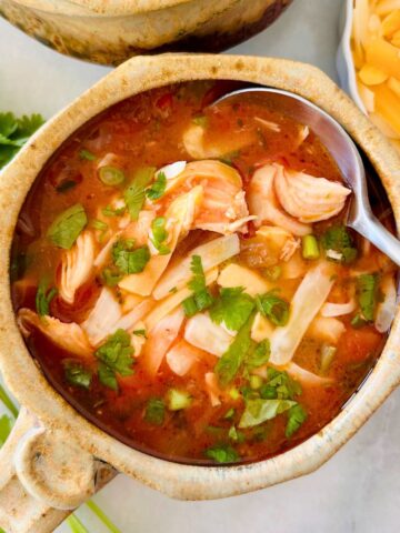 A spoon is dipping into a potted mug of Mexican chicken and vegetable soup sitting between cilantro sprigs and shredded cheese.
