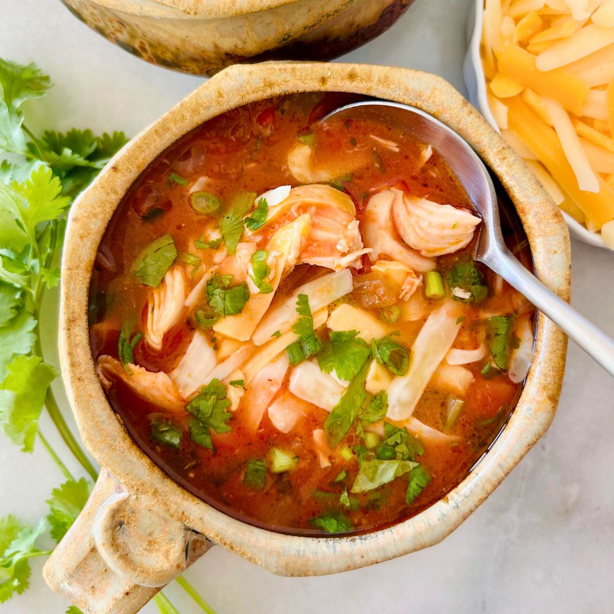 A spoon is dipping into a potted mug of Mexican chicken and vegetable soup sitting between cilantro sprigs and shredded cheese.